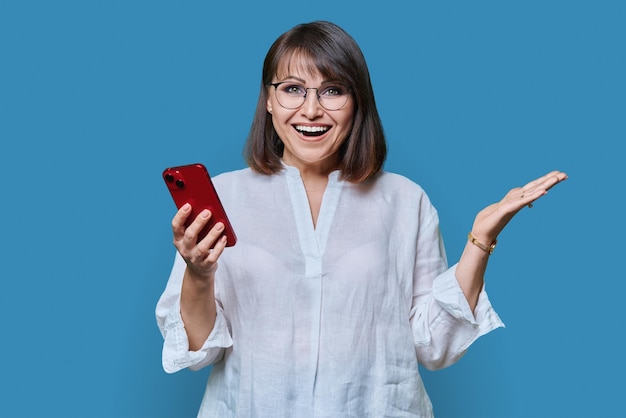 Retrato de mujer de mediana edad con teléfono mirando a la cámara sobre fondo azul.