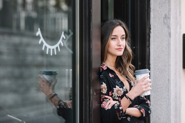 Retrato de mujer de moda con café para ir apoyándose en la puerta de entrada de su tienda