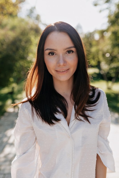 Retrato de mujer de moda con camisa blanca y cabello oscuro sonríe a la cámara en la calle a la luz del sol Adorable dama bonita pasa tiempo afuera en la ciudad