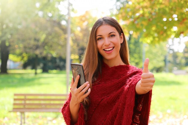 Retrato de mujer de moda con teléfono inteligente mostrando el pulgar hacia arriba en el parque de la ciudad.