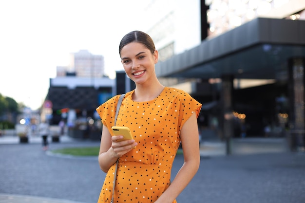 Retrato mujer de moda con vestido amarillo caminando por la calle y sosteniendo el teléfono inteligente en la mano