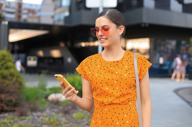 Retrato mujer de moda en vestido amarillo con gafas de sol caminando por la calle y sosteniendo el teléfono inteligente en la mano