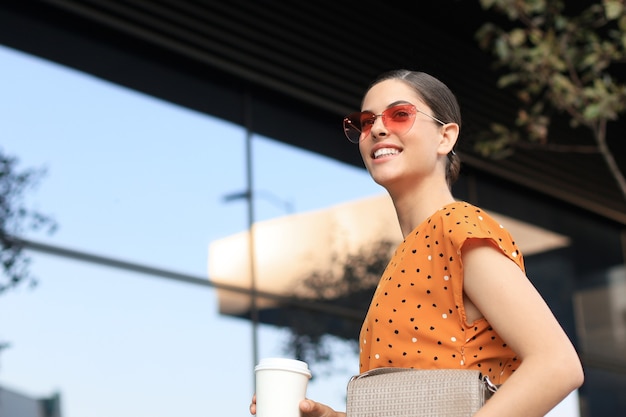 Retrato de mujer de moda en vestido amarillo con taza de café caminando por la calle de la ciudad, sonriendo al lado.