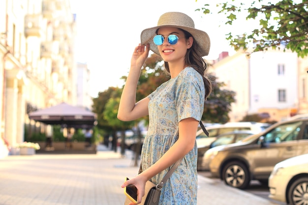 Retrato de mujer de moda en vestido azul con gafas de sol caminando en la calle y sosteniendo el teléfono inteligente en la mano.