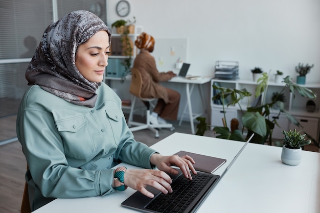 Retrato de mujer moderna del Medio Oriente vistiendo velo en la oficina y usando la computadora portátil, espacio de copia
