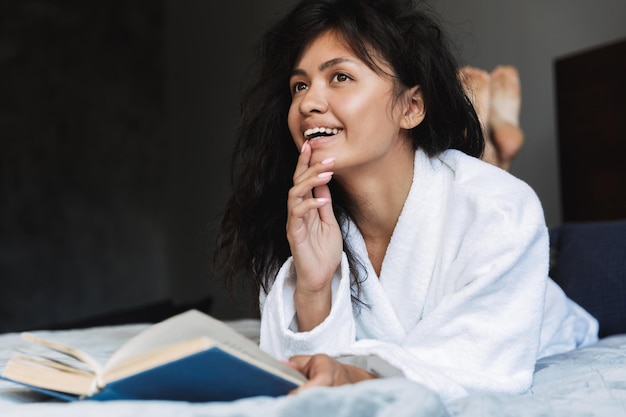 Retrato de mujer morena alegre vistiendo bata libro de lectura en la cama en casa