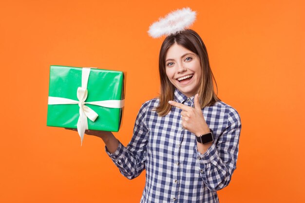 Retrato de una mujer morena angelical positiva con una sonrisa encantadora usando un pantalones a cuadros de pie con halo en la cabeza y apuntando a una caja de regalo, caridad. tiro de estudio interior aislado sobre fondo naranja