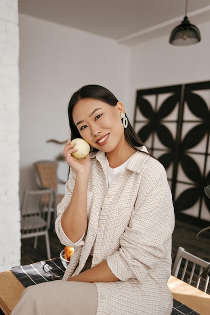 Retrato de mujer morena con chaqueta y pantalones beige sonríe ampliamente y se sienta en una mesa de madera en la cocina Encantadora chica asiática de buen humor sostiene manzana