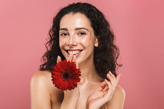 Retrato de mujer morena complacida de 20 años con cabello largo sonriendo y sosteniendo una flor roja, aislada sobre pared rosa