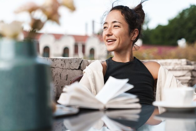 Foto retrato de mujer morena complacida en ropa casual leyendo un libro y tomando café mientras descansa en la terraza al aire libre