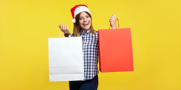 Retrato de una mujer morena compradora satisfecha con un sombrero de santa de año nuevo y una sonrisa encantadora de pie mostrando bolsas de compras con la compra a la cámara en un estudio interior aislado en un fondo amarillo