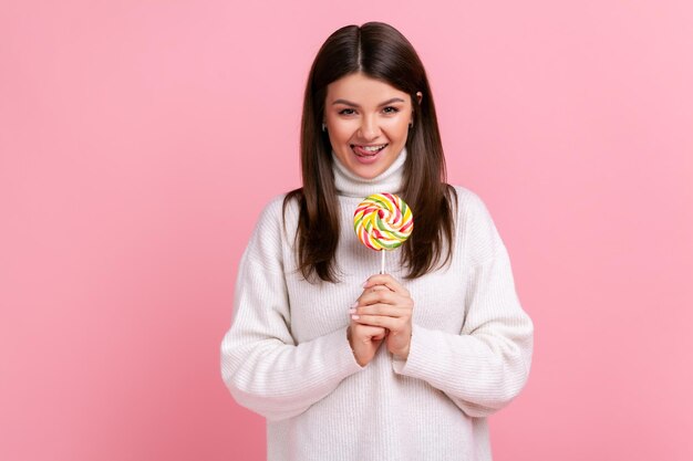 Retrato de mujer morena hambrienta sosteniendo grandes dulces sabrosos, mirando a la cámara, mostrando la lengua, usando un suéter blanco de estilo casual. Disparo de estudio interior aislado sobre fondo rosa.