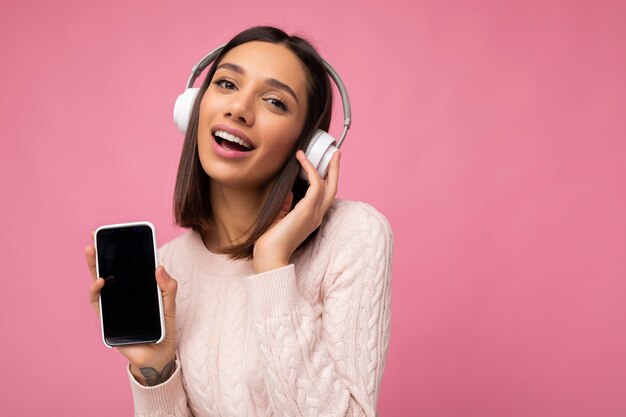 Retrato de mujer morena joven linda hermosa sexy con suéter rosa aislado sobre la pared de fondo rosa con auriculares inalámbricos bluetooth blancos y escuchando buena música y mostrando el móvil