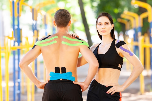 Retrato de mujer morena con la mano en el hombro del hombre irreconocible, atletas caucásicos profesionales con cinta kinesiológica en los cuerpos, posando en el campo de deportes, mirando a la cámara.