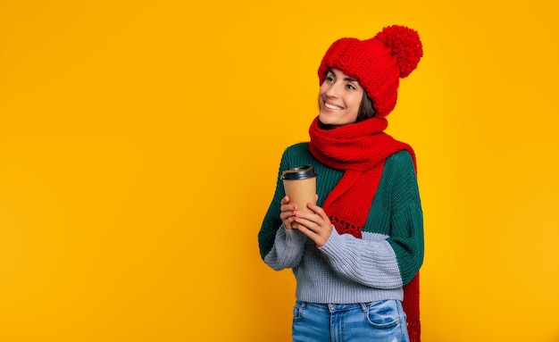 Retrato de mujer morena moderna feliz con sombrero rojo y bufanda de cerca mientras posa con una taza de bebida en la mano en la pared amarilla en estudio