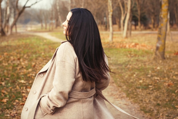 Retrato de mujer morena de pelo en abrigo beige caminando en el parque de la ciudad