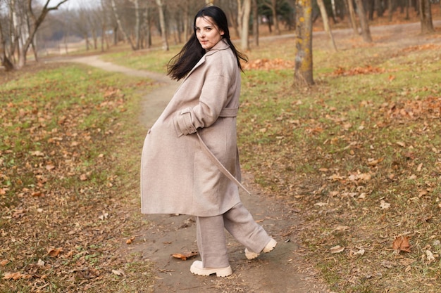 Retrato de mujer morena de pelo en abrigo beige caminando en el parque de la ciudad