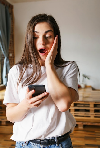 Retrato de mujer morena de pelo largo con camiseta blanca mirando su teléfono inteligente con cara sorprendida