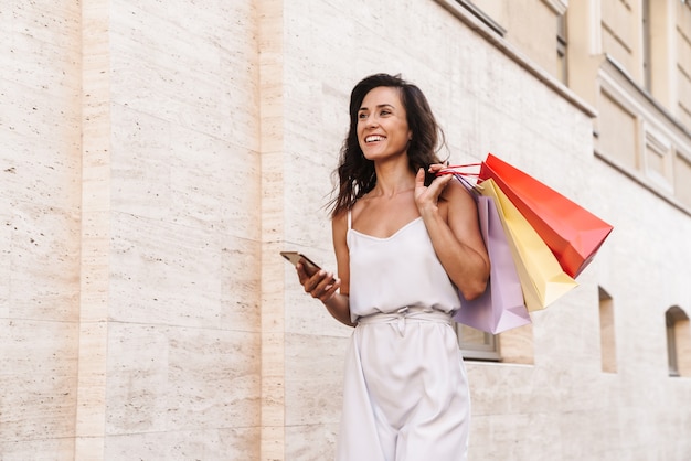 Retrato de mujer morena sonriente con teléfono celular y sosteniendo coloridas bolsas de compras mientras camina por las calles de la ciudad