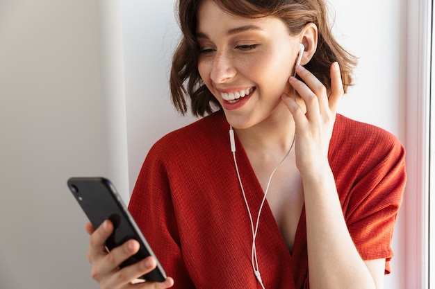 Retrato de mujer morena sonriente vestida con ropa casual escuchando música con teléfono inteligente y auriculares mientras está sentado sobre la ventana en casa