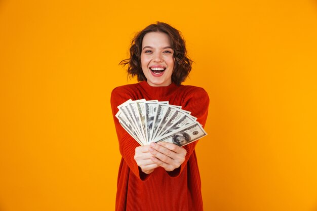 Retrato de mujer morena vistiendo suéter sosteniendo un montón de dinero en efectivo mientras está de pie aislado en amarillo