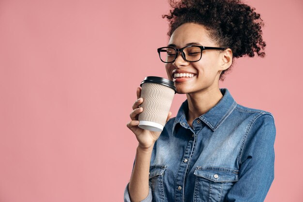 Foto retrato de la mujer multirracial alegre y rizada que sostiene una taza de papel y huele a café para aislarse en rosa