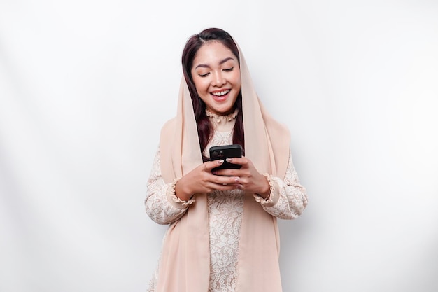 Un retrato de una mujer musulmana asiática feliz con un pañuelo en la cabeza sosteniendo su teléfono aislado por un fondo blanco