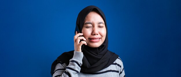 Foto retrato de mujer musulmana asiática recibe malas noticias en el teléfono, expresión de llanto triste. sobre fondo azul