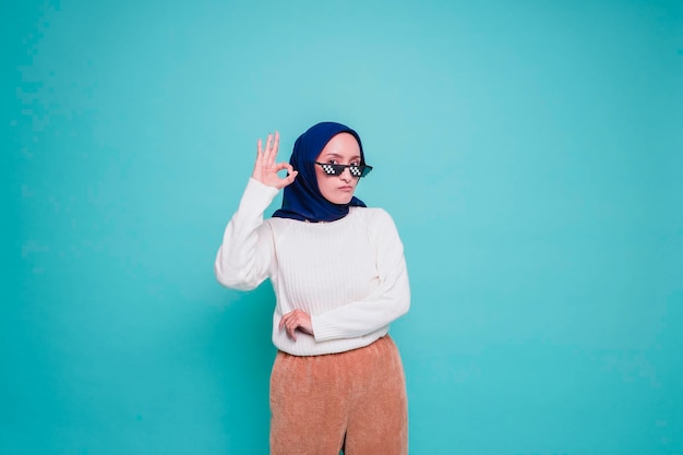 Retrato de una mujer musulmana asiática sonriente con gafas dando un gesto de mano OK aislado sobre fondo azul.