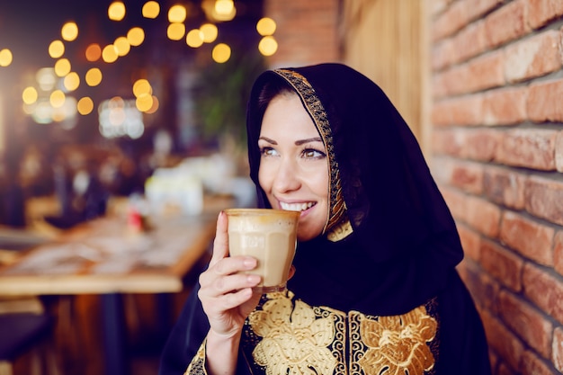 Retrato de mujer musulmana impresionante en ropa tradicional sentado en la cafetería y tomando café.