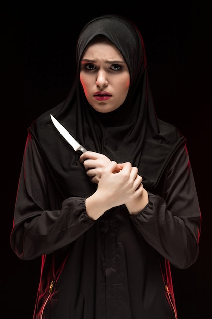 Retrato de una mujer musulmana joven asustada asustada desesperada hermosa que lleva el hijab negro que sostiene el cuchillo en sus manos