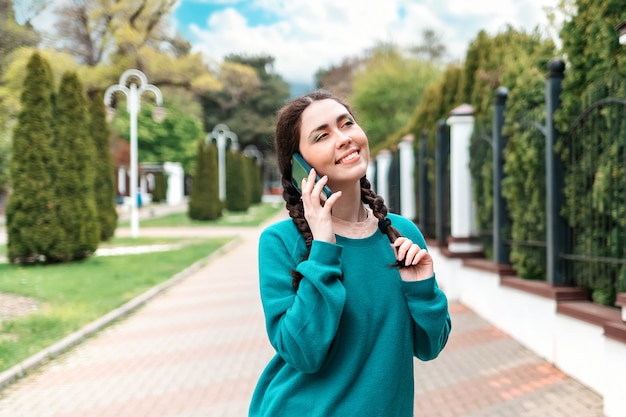 Retrato de una mujer muy sonriente con coletas hablando por teléfono. Al fondo hay un callejón con árboles. Concepto de comunicación.