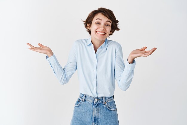 Retrato de una mujer natural feliz encogiéndose de hombros y sonriendo sin un gran gesto de pie despreocupado sobre fondo blanco