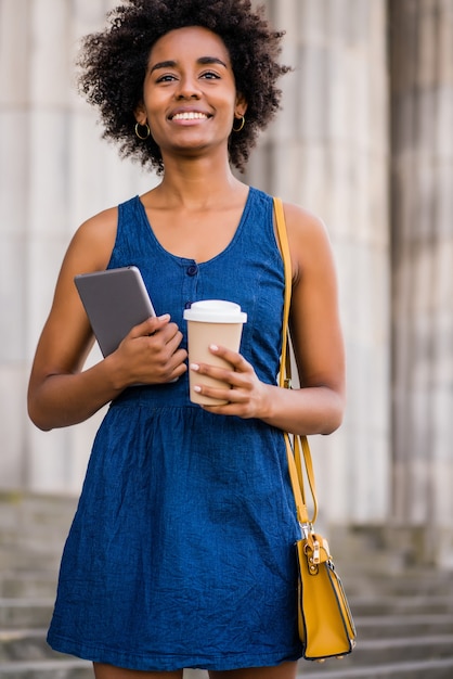 Retrato de mujer de negocios afro sosteniendo una tableta digital y una taza de café mientras está parado al aire libre en la calle. Concepto urbano y empresarial.