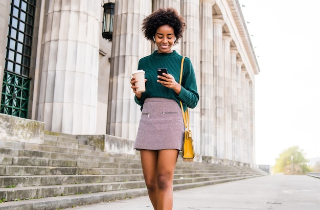 Retrato de mujer de negocios afro usando su teléfono móvil y sosteniendo una taza de café mientras camina al aire libre en la calle. Concepto urbano y empresarial.
