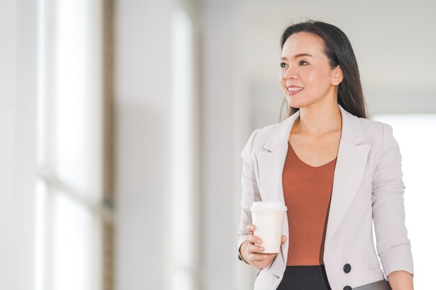 Retrato de mujer de negocios asiática alegre sostiene café para llevar y tableta digital en la empresa