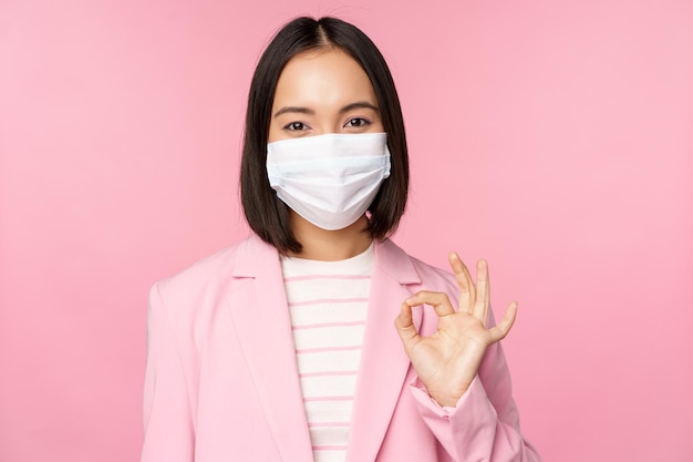 Retrato de una mujer de negocios asiática con mascarilla médica que muestra un signo de estar bien usando reglas de trabajo de traje durante la pandemia de covid19 de pie sobre fondo rosa