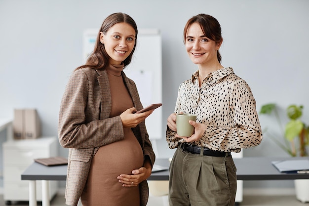 Retrato de mujer de negocios embarazada con teléfono móvil sonriendo a la cámara junto con su colega con una taza de café que están de pie en la oficina