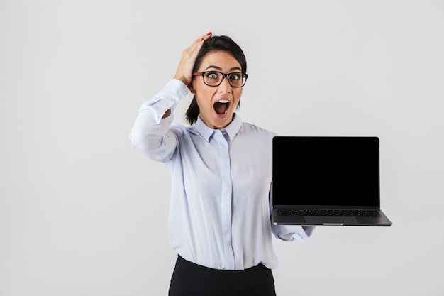 Retrato de mujer de negocios emocionada con anteojos sosteniendo un portátil plateado en la oficina, aislado sobre la pared blanca