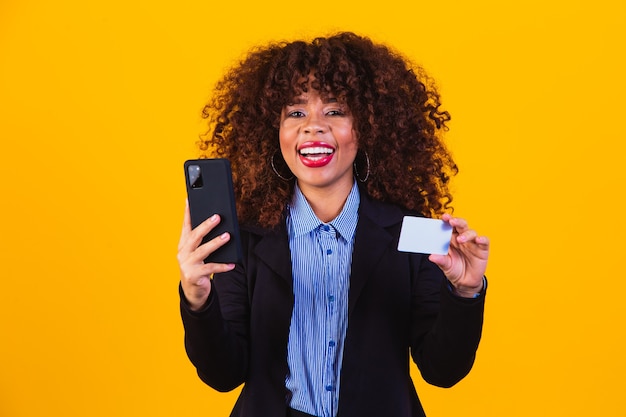 Retrato de mujer de negocios emocionada mirando el teléfono celular mientras está de pie y sosteniendo la tarjeta de crédito aislada sobre fondo amarillo.