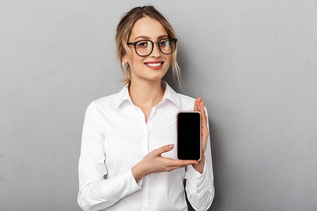 Retrato de mujer de negocios europea con gafas sonriendo y sosteniendo el teléfono inteligente en la oficina, aislado