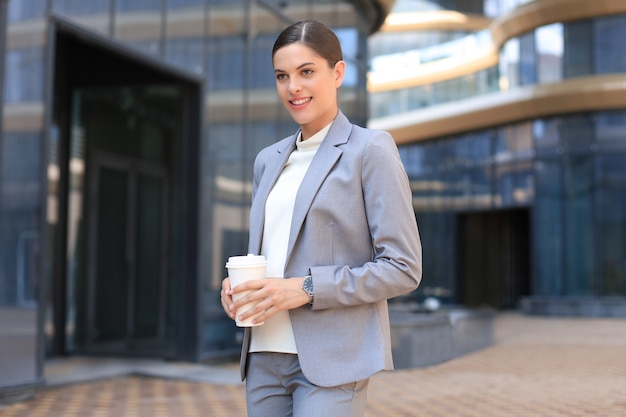 Retrato de mujer de negocios exitosa que va a trabajar con café caminando cerca del edificio de oficinas en la calle de la ciudad.