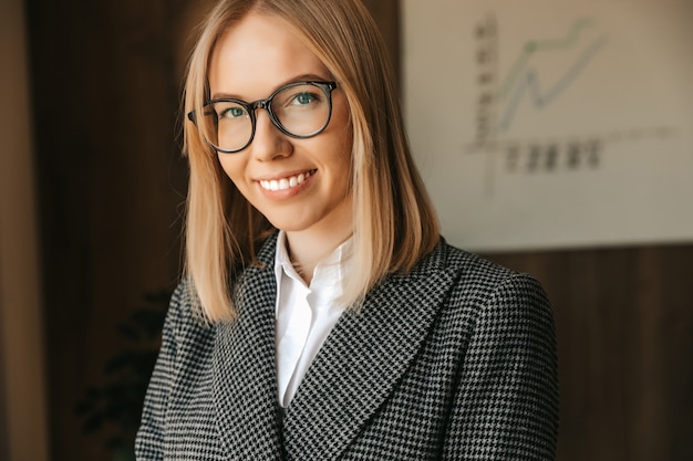 Retrato de una mujer de negocios con gafas, en un estricto traje de negocios, sonriendo con dientes en la oficina. Trabajador feliz exitoso de oficina.