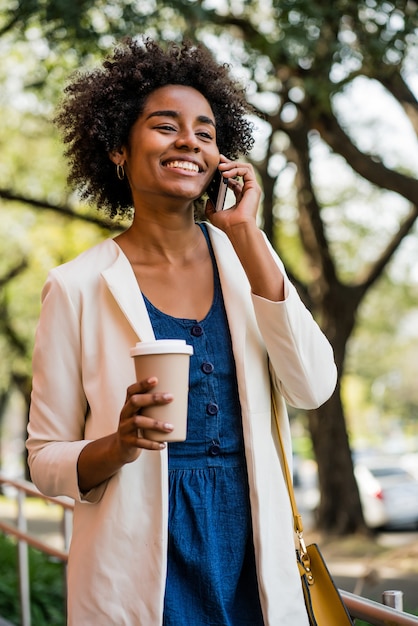 Retrato de mujer de negocios hablando por teléfono y sosteniendo una taza de café mientras está de pie al aire libre en el parque. Concepto de negocio.