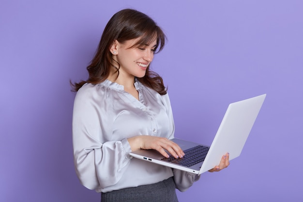 Retrato de mujer de negocios joven alegre que trabaja en la computadora portátil y sonriente, mujer con computadora portátil en las manos, feliz, vistiendo blusa y falda, dama atractiva navegando por Internet para trabajar.