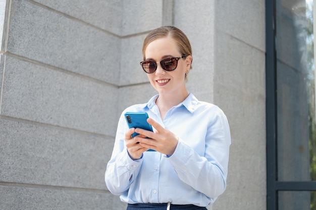 Retrato de mujer de negocios joven confiada feliz en sunglasess, escribiendo en el teléfono mientras está parado en la ciudad al aire libre. Foto de alta calidad