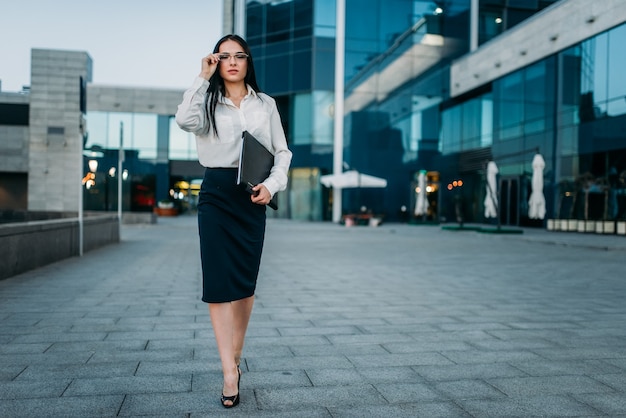 Retrato de mujer de negocios joven con gafas y blusa blanca con cuaderno en mano, skyscaper. Edificio moderno, centro financiero, paisaje urbano.