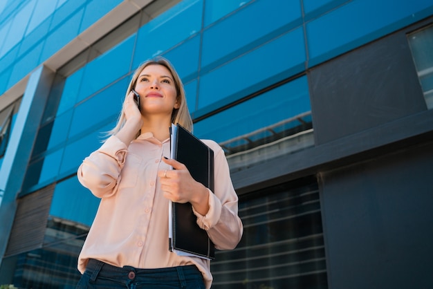 Retrato de mujer de negocios joven hablando por teléfono mientras está de pie fuera de los edificios de oficinas