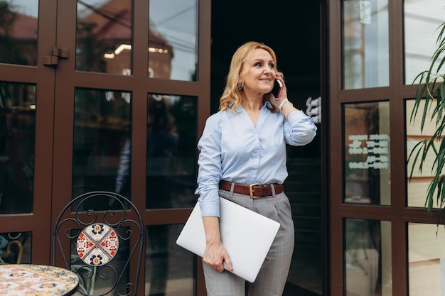 Retrato de una mujer de negocios madura feliz usando su teléfono mientras está de pie en la acera de un café