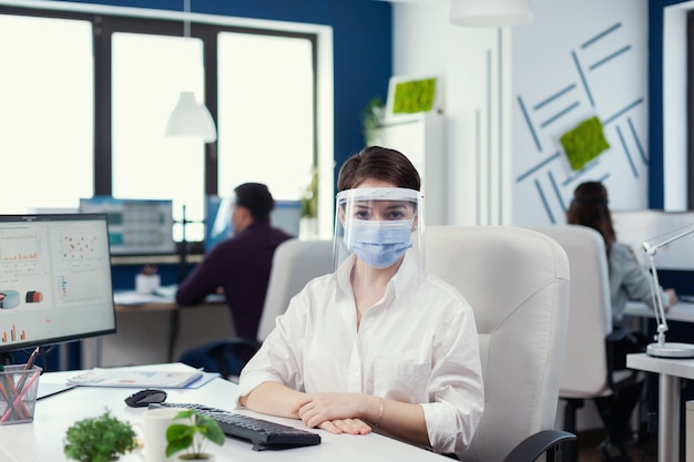 Retrato de mujer de negocios mirando a la cámara sentado en una silla con máscara facial contra covid19. Equipo empresarial que trabaja en una empresa financiera respetando la distancia social durante la pandemia global.